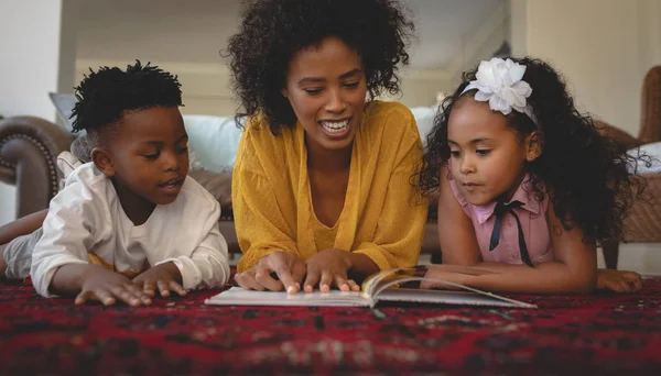 彼女のかわいい子供たちが床の上に横たわると 快適な家で絵本を読んでとアフリカ系アメリカ人の母の正面図 — ストック写真
