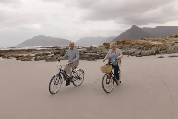 在背景为山的海滩上骑自行车的活跃的高级夫妇的正面看法 — 图库照片