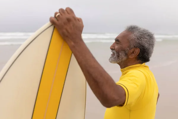 ビーチにサーフボードを抱えて立っているハッピー シニア男性サーファーの側面図 — ストック写真
