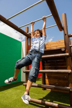 Low angle view of a mixed-race schoolboy playing on horizontal ladder in the school playground on a sunny day clipart