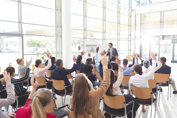 Rear view of interactive diverse business people listening to Caucasian businessman and raising hands to ask him questions in conference