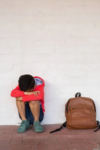小学校の廊下の床に彼の隣に彼の袋のパックで一人で座って悲しい混血少年の正面図 — ストック写真