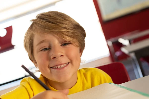 小学校の教室の机で本にスケッチを描きながらカメラを見て幸せな白人少年の正面図 — ストック写真