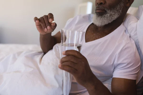 在家里的卧室里 非洲裔美国高级男子中段用一杯水服药 — 图库照片