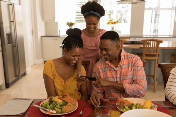 自宅のダイニング テーブルに座って 娘の携帯電話を見て幸せのアフリカ系アメリカ人の両親の正面図 — ストック写真