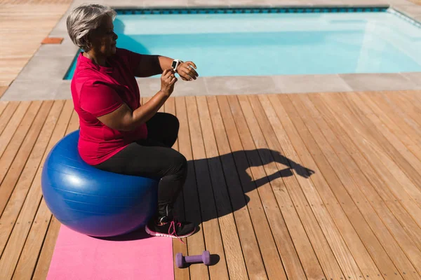 家の裏庭にプールの横にある運動ボールのスマートウォッチを使用してアフリカ系アメリカ人のアクティブなシニア女性の側面図 — ストック写真