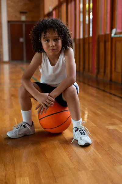 学校篮球场上坐在篮球上看镜头的混血小学生的正面 — 图库照片