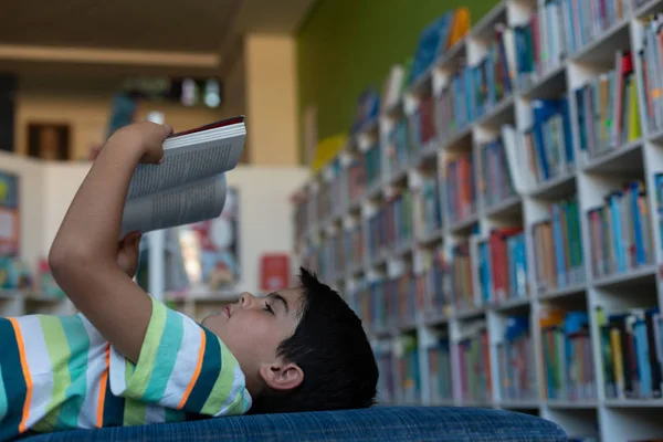 ソファーで横になっていると 小学校の図書館で本を読んで白人少年の側面図 — ストック写真