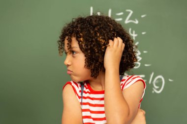 Front view of a thoughtful mixed-race schoolboy holding his head and standing against the green board in a classroom at school clipart