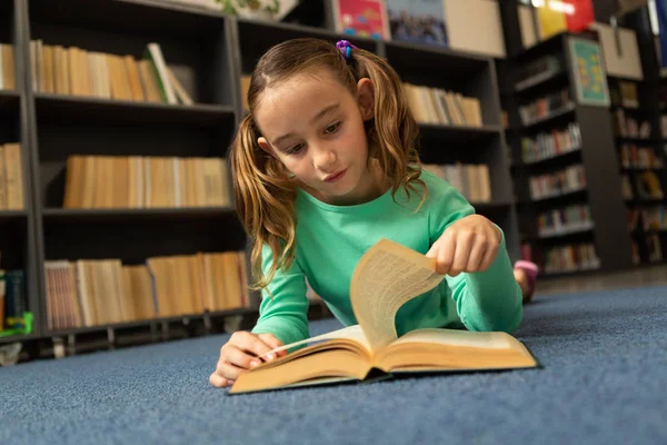 床の上に横たわると 小学校の図書館の本のページをめくる白人女子高生の正面図 — ストック写真