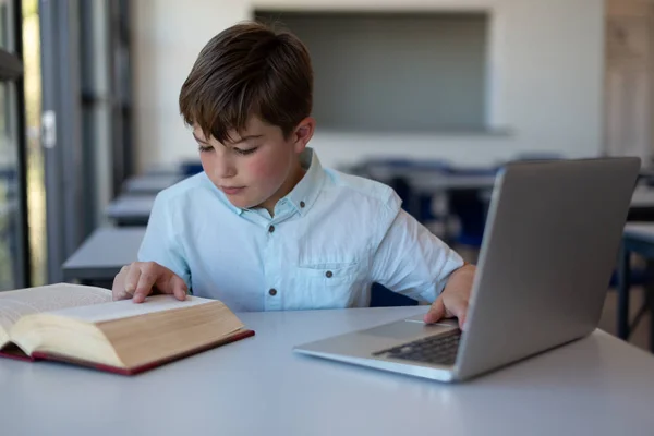 在学校的一间教室里 一个高加索小学生一边用笔记本电脑看书 一边看前面的风景 — 图库照片