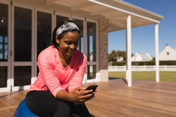 家の裏庭にエクササイズ ボールに座りながら携帯電話を使用してアフリカ系アメリカ人の中年の女性の正面図 — ストック写真