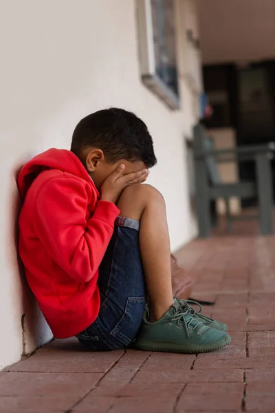 小学校の廊下の床に一人で座って悲しい混血少年の側面図 — ストック写真