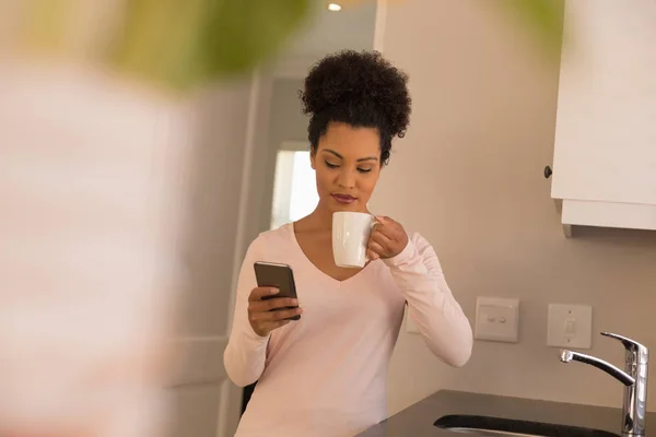 非洲裔美国人美女在家里的厨房里使用手机时喝咖啡的正面看法 — 图库照片
