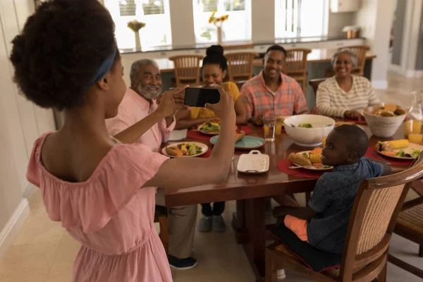 アフリカ系アメリカ人の女の子が自宅彼女の携帯電話をダイニング テーブルの周り彼女の家族の写真を撮るの背面図 — ストック写真