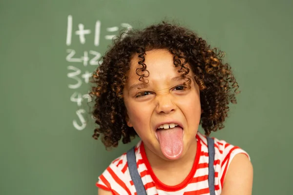 緑の基板に対して立っていると学校で教室でカメラで舌を抜くかなり混血少年の正面図 — ストック写真