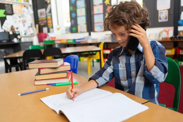 学校の教室の机に自分のノートに書きながら携帯電話で話している白人少年の側面図 — ストック写真