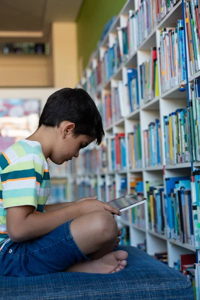 在小学的图书馆里 一名白种人小学生坐在图书馆的沙发上 一边使用数字平板电脑的侧视图 — 图库照片