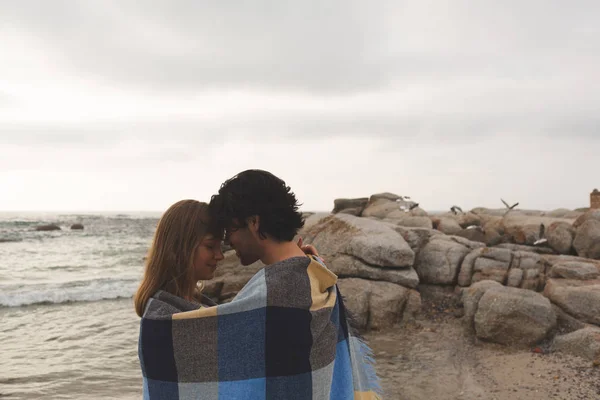 站在海滩上的年轻的高加索夫妇在毯子里互相亲吻的侧视图 — 图库照片