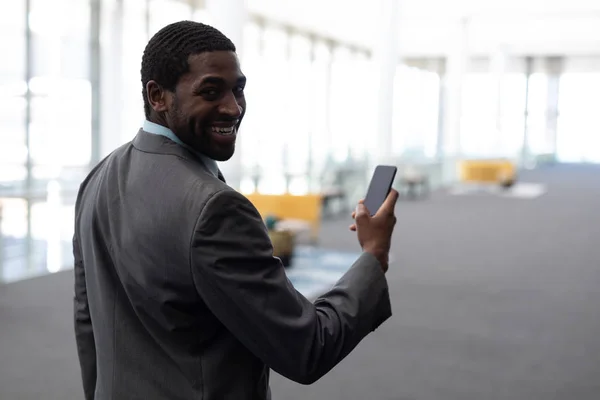 近代的なオフィスに立っている携帯電話を使用して幸せな若いアフリカ系アメリカ人実業家の側面図です 彼は見て カメラに笑顔 — ストック写真