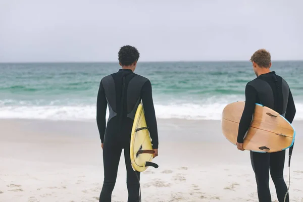 サーフボードを押しながらビーチに立っている若い多民族の男性の友人の背面図 — ストック写真