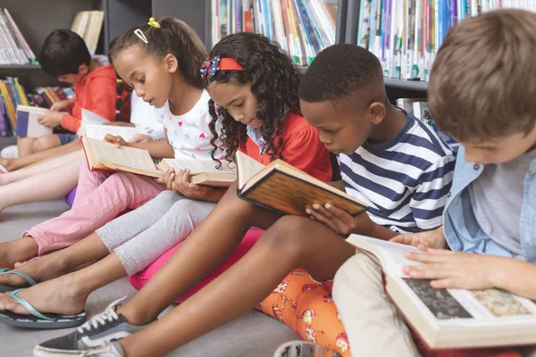 ミックス民族性スクールの子供たちの側面は クッションの上に座って バックグラウンドで Bookshelfs に対して学校で図書館で本の上に勉強します — ストック写真