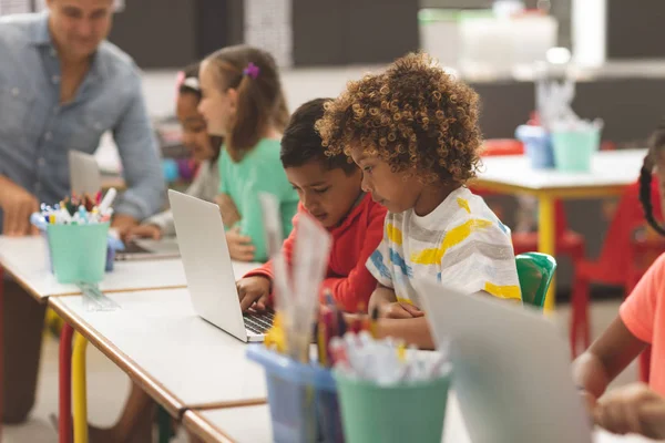 教師が学校でのバックグラウンドで教えている間 教室でノートパソコンを使用して 学校の子供の側面図 — ストック写真