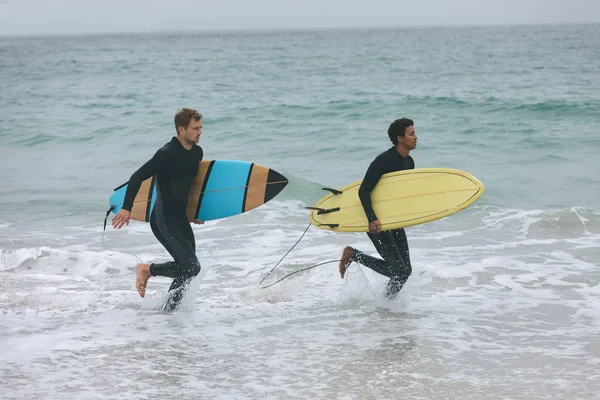 サーフボードを押しながらビーチを走る若い多民族の男性の友人の側面図 — ストック写真