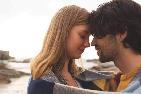 站在海滩上的年轻的高加索夫妇在毯子里互相亲吻的侧视图 — 图库照片