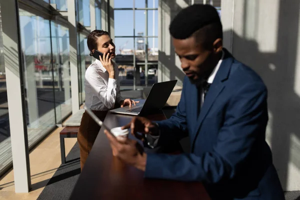 在现代办公室使用笔记本电脑时 年轻的高加索女商人一边用手机说话 在背景中使用数字平板电脑的非裔美国人同事 — 图库照片