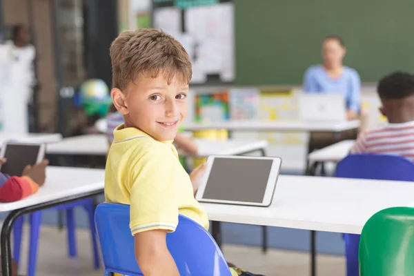 学校の教室で カメラを見て 机の上にデジタルタブレットを持っている幸せな男子生徒の背面図 — ストック写真