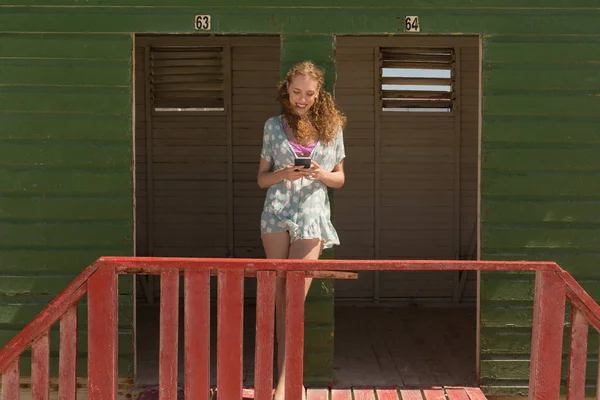 在海滩小屋使用手机的快乐华丽的年轻白人妇女的前面的看法 她在微笑 — 图库照片
