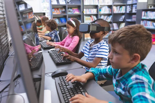 Side view of African-american schoolboy using virtual reality headset while his schoolmates working on computer  in computer room