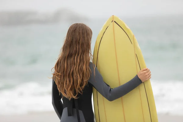 ビーチにサーフボードを抱えて立っている女性サーファーの背面図 — ストック写真