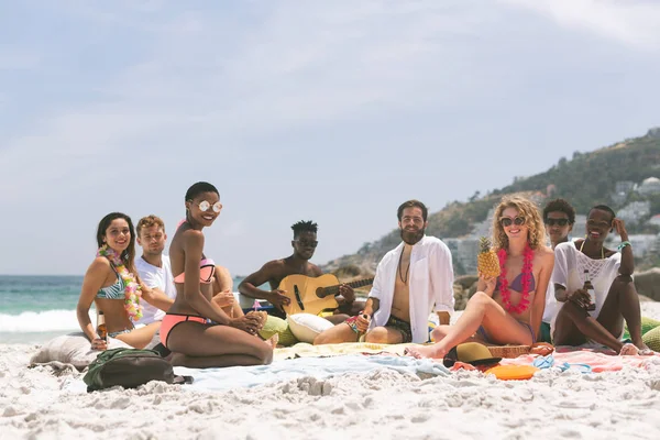 Front view of multi ethnic group of friends relaxing and sitting on beach while looking at camera