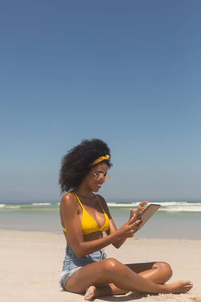 黄色ビキニ ビーチに座ってデジタル タブレットを使用してサングラスの若いアフリカ系アメリカ人女性の側面図です 彼女は笑っています — ストック写真