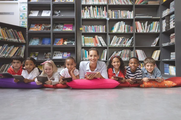 バックグラウンドで Bookshelfs に対して図書館で彼の学校の子供たちに横たわっている幸せな教師の表面レベルのビュー — ストック写真