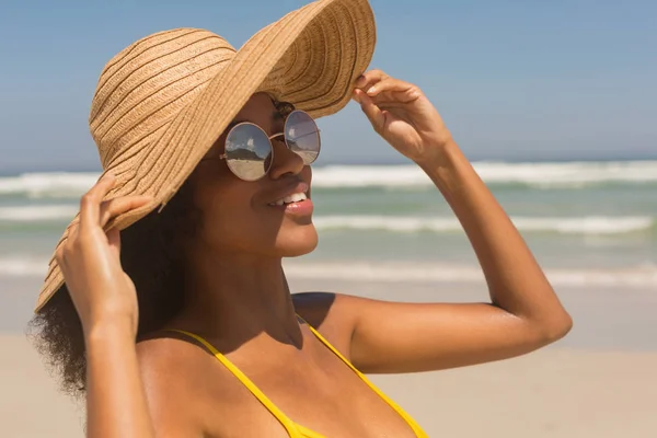 穿着黄色比基尼 帽子和太阳镜的年轻非洲裔美国妇女在阳光下站在沙滩上的特写镜头 她在微笑 — 图库照片