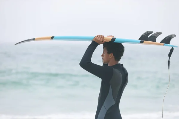 ビーチで彼女の頭の上にサーフボードを運ぶ若い混血男性サーファーの側面図 — ストック写真