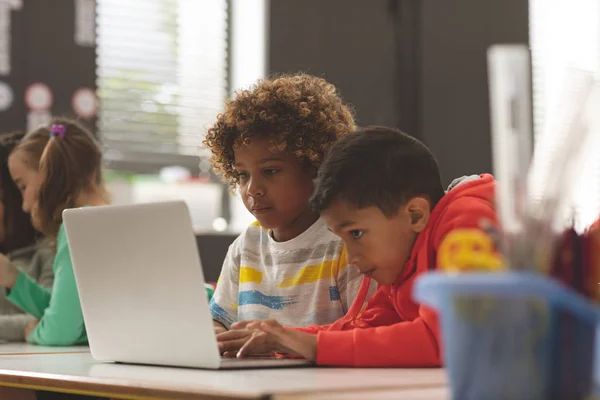 两个学校的孩子在教室里工作的一个笔记本电脑对背景的学校女孩的前面的看法 — 图库照片