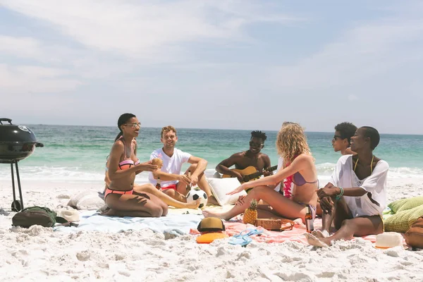 front view of multi ethnic group of friends relaxing and sitting on beach while interacting with each other. They are sitting on beach towel, African-american playing guitar