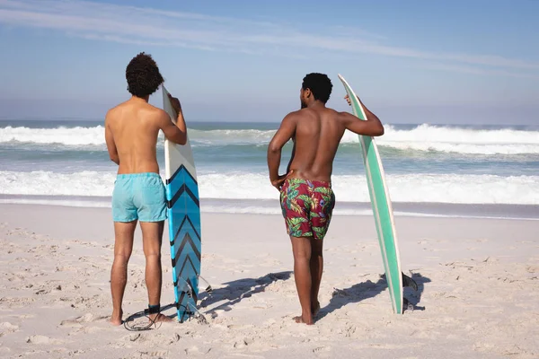太陽の下で浜辺の海の前に立っている間にサーフボードを持っている若い多民族男性の背面図 — ストック写真