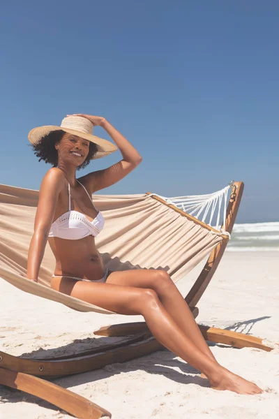 晴れた日にビーチでカメラに笑顔ながらハンモックに座って帽子の白ビキニの若い美しい混血の女性の肖像画 — ストック写真