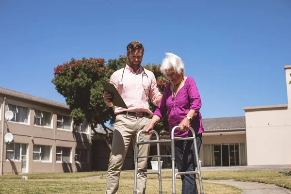 一个白人男医生帮助高级白人妇女与她的步行者在后院养老院的低视角 — 图库照片