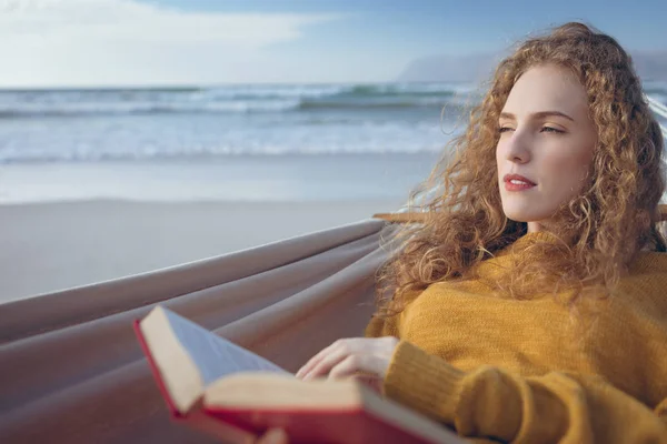 在阳光明媚的日子里 在沙滩上躺在吊床上的时候 穿着漂亮的高加索妇女的书的正面景色 她把目光移开 — 图库照片