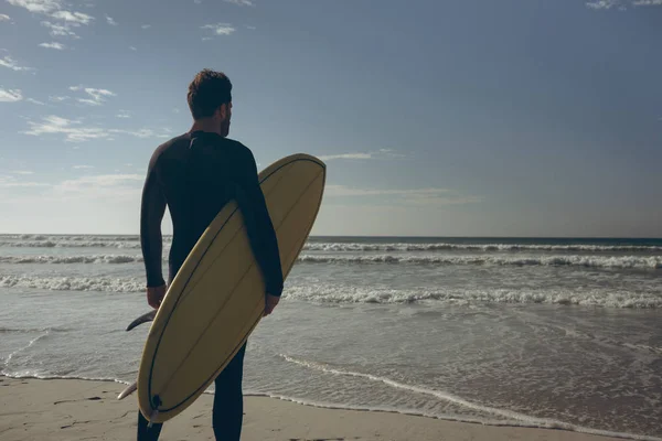 晴れた日のビーチで海にサーフボードに立って顔をウェット スーツの若い白人男性サーファーの後姿 彼は彼のサーフボードを抱えています — ストック写真