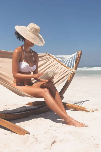 在阳光明媚的日子里 穿着白色比基尼的年轻漂亮混血女子坐在沙滩上的吊床上 用数字平板电脑戴着帽子 可以看到前厅的景色 — 图库照片