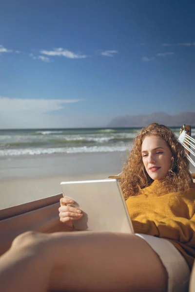 阳光明媚的日子里 美丽的高加索妇女躺在沙滩上的吊床上 使用数字平板电脑欣赏美丽的高加索妇女的美景 — 图库照片