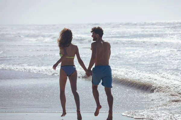 在阳光明媚的日子里 在海滩上 快乐的年轻高加索夫妇牵手在海边奔跑 他们在微笑 — 图库照片