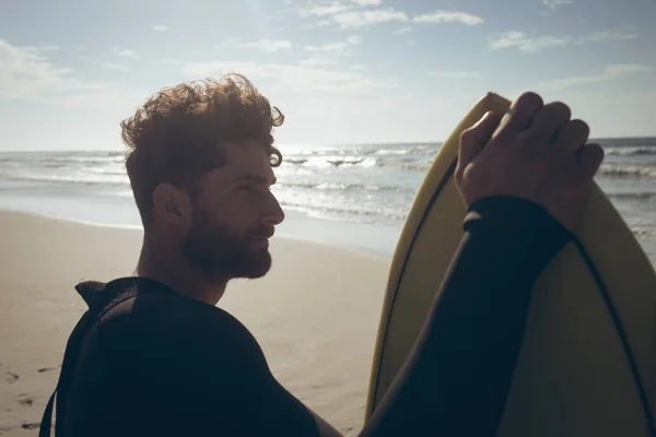 在阳光明媚的日子里 年轻的高加索男性冲浪者的侧视图与冲浪板站在海滩上 他把目光移开 — 图库照片